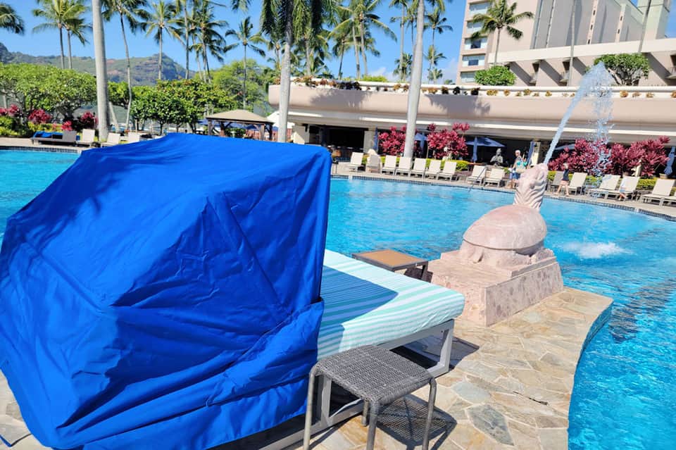 Resort pool area with blue pop-up shade tent, decorative sculpture, and palm-lined gardens