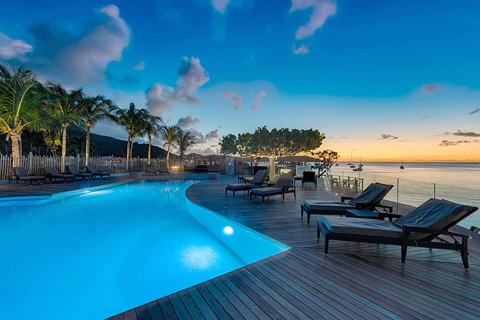 Illuminated infinity pool overlooking ocean at dusk with lounge chairs and palm trees