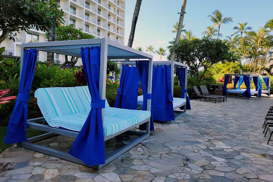 Contemporary poolside cabanas with blue curtains, striped cushions, tropical plants, and hotel building in background