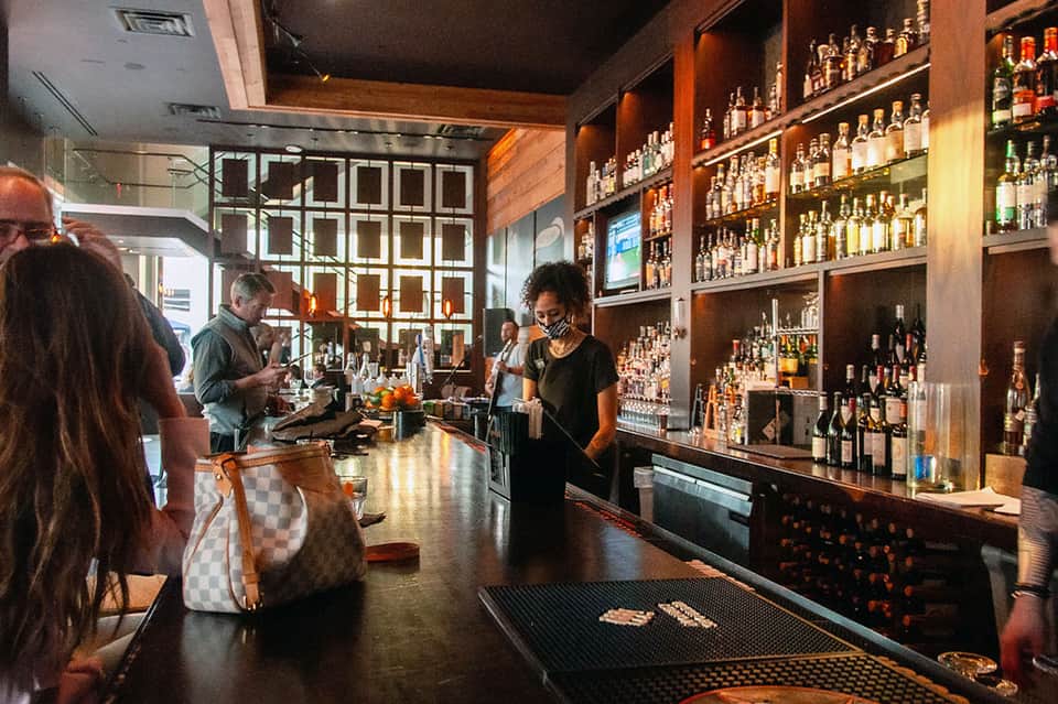 Modern bar interior with extensive bottle collection, wood accents, geometric wall design, and bartender serving customers