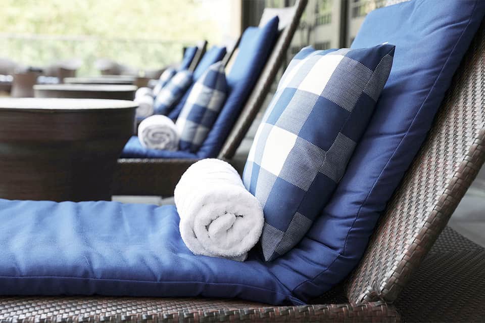 Outdoor lounge seating with blue cushions, checkered pillows, and rolled white towels on wicker furniture