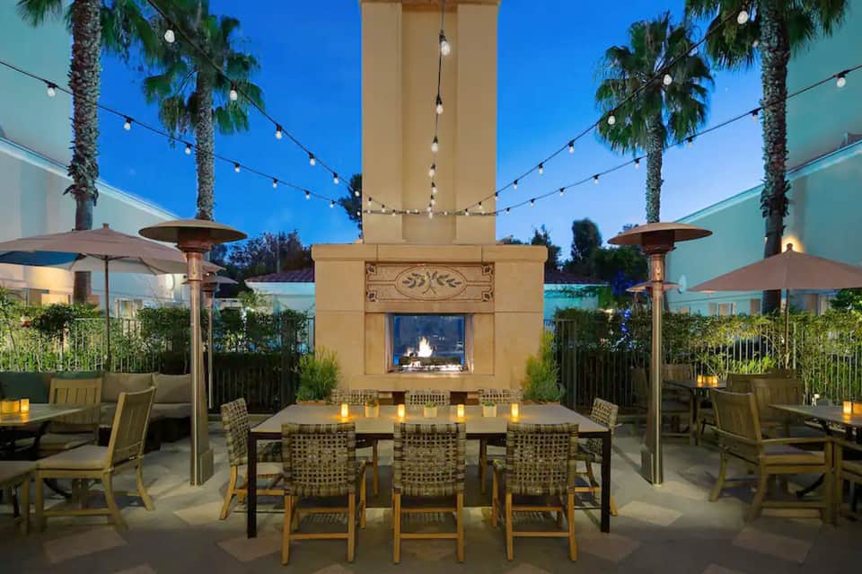 Elegant outdoor dining terrace with fireplace, string lights, palm trees, and formal table setting at dusk