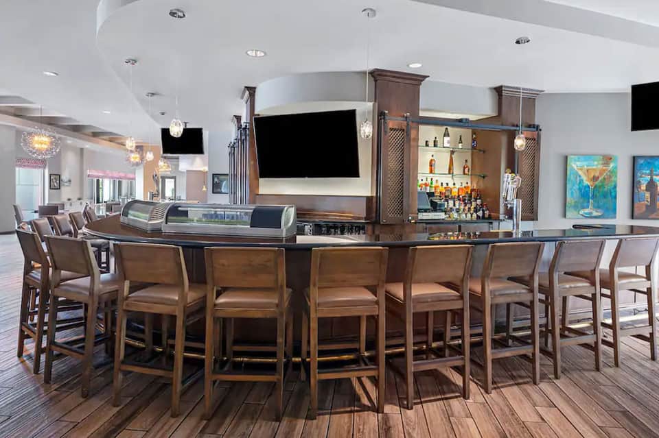 Hotel bar with curved counter, wood accents, television screens, and pendant lighting