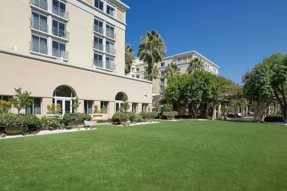 Modern hotel courtyard with manicured lawn, palm trees, arched architecture, and tall residential building