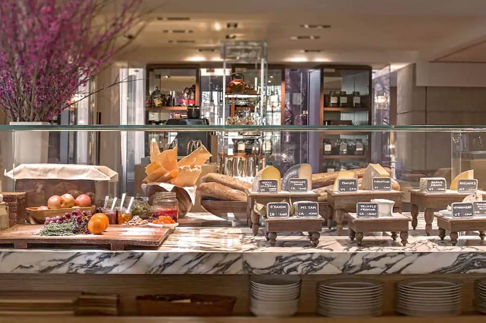 Upscale bakery display counter with fresh bread, pastries, fruits, and labeled items under warm lighting