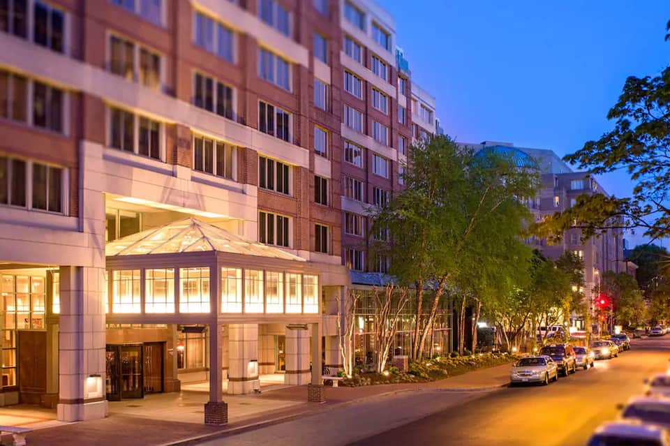 Modern hotel entrance with illuminated glass canopy at dusk on a tree-lined urban street