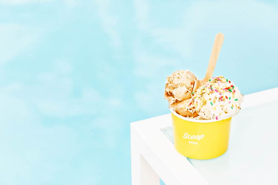 Yellow ice cream cup with two scoops and colorful sprinkles on wooden spoon against bright blue sky