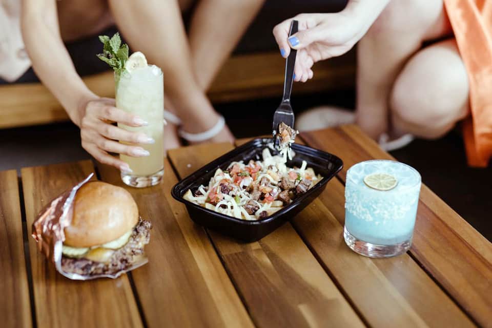 Hands holding cocktails and sharing appetizers on wooden table at waterfront restaurant