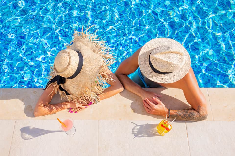 Two people relaxing by pool wearing straw hats with drinks and towels poolside