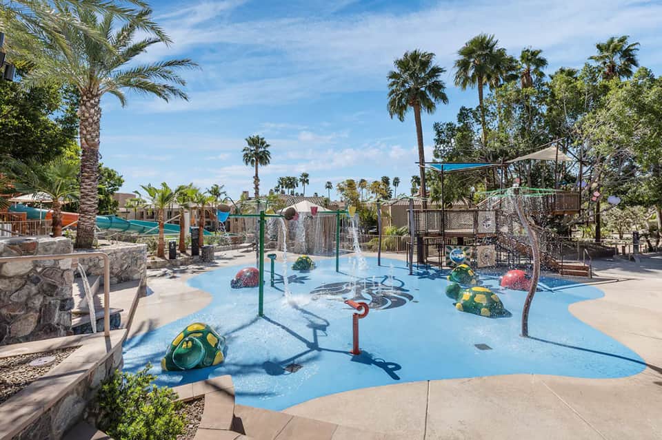 Colorful splash pad playground with water features, palm trees, and desert landscape