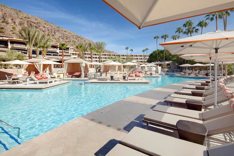 Resort pool with lounge chairs and orange umbrellas beneath palm trees and desert mountain