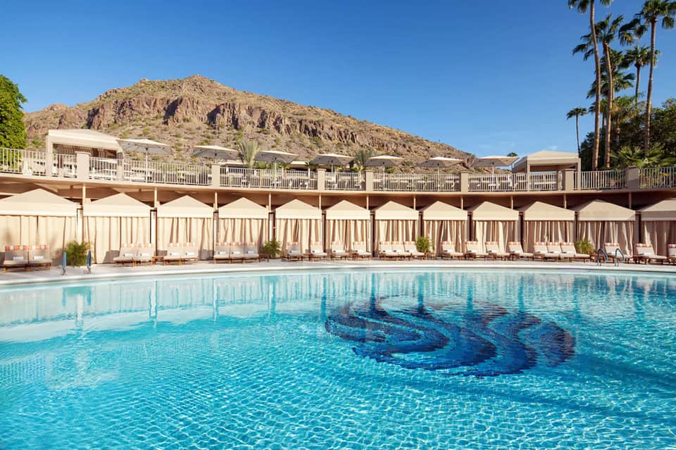 Resort pool with turquoise water, beige cabanas with umbrellas, palm trees, and rocky mountain backdrop during daytime