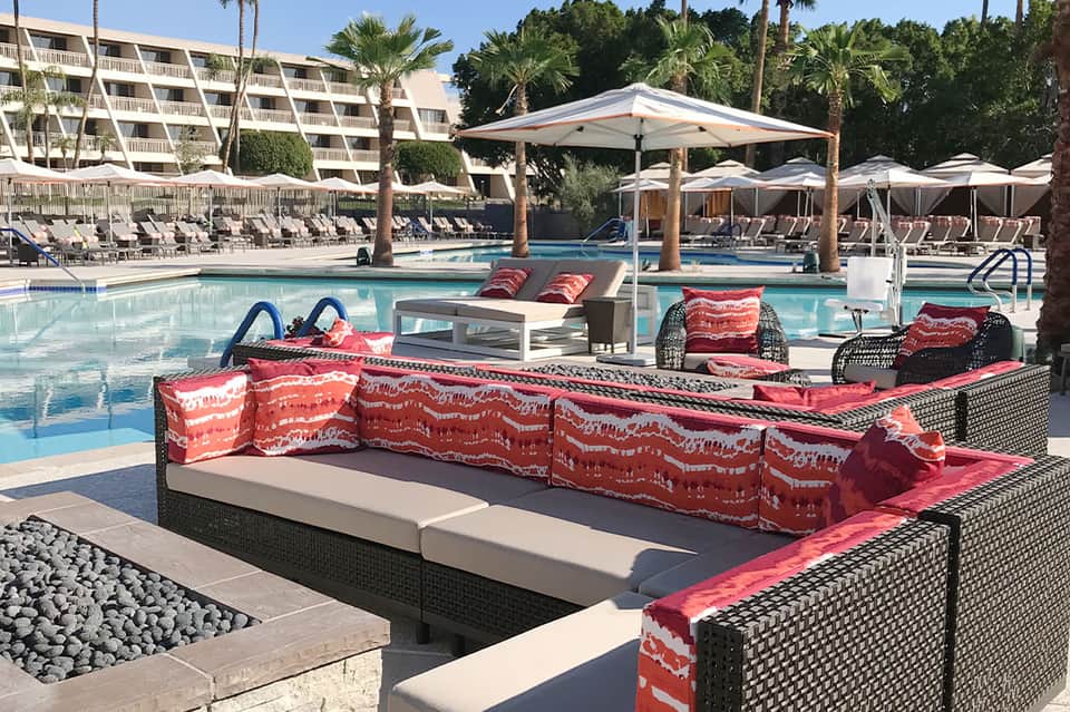 Resort pool deck with red patterned loungers, umbrellas, palm trees, and residential buildings