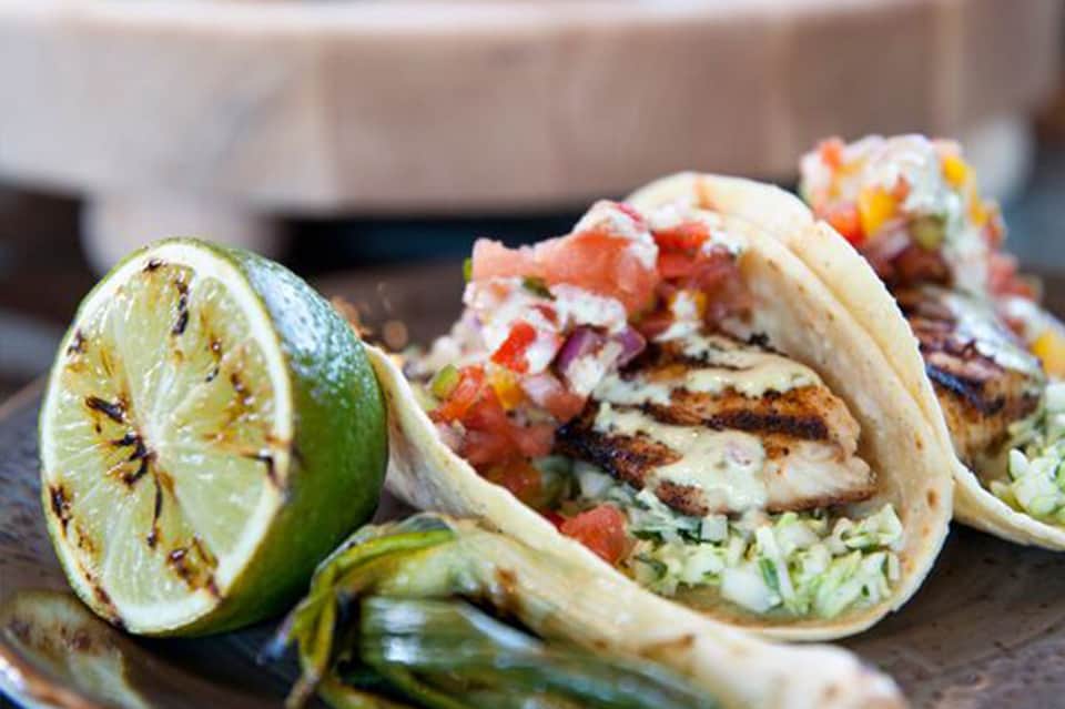 Grilled fish tacos with fresh lime and colorful toppings on wooden surface