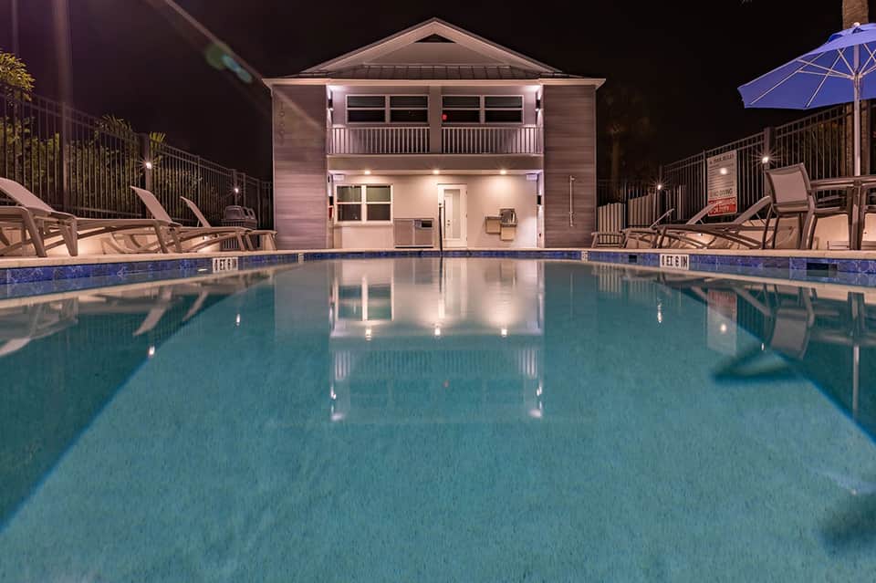 Illuminated resort pool at night with two-story pool house, lounge chairs, blue umbrella, and deck lighting