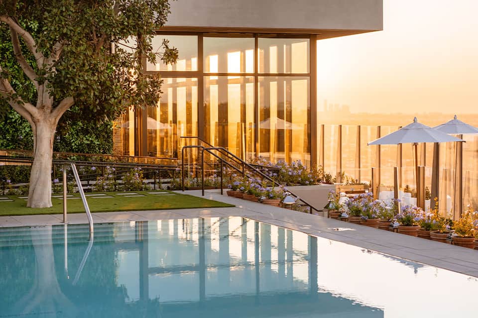 Infinity pool at sunset with modern building, tree, and blue umbrellas overlooking valley