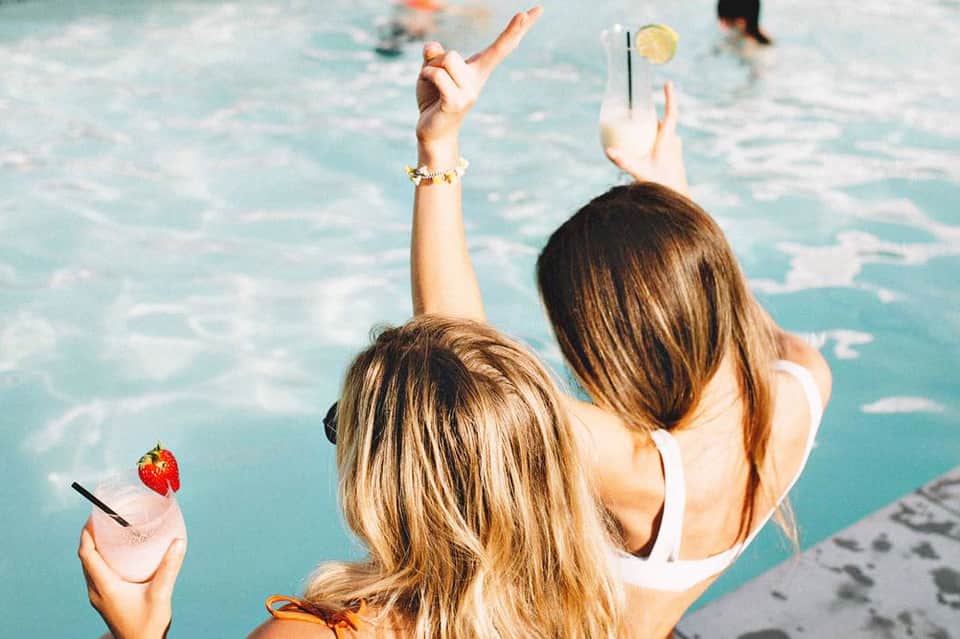 Two women at poolside holding tropical cocktails with straws and fruit garnishes