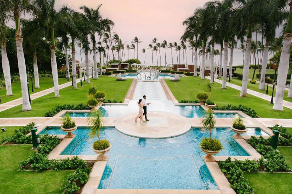 Luxury resort grounds with palm-lined pools, fountains, manicured lawn, and ocean view