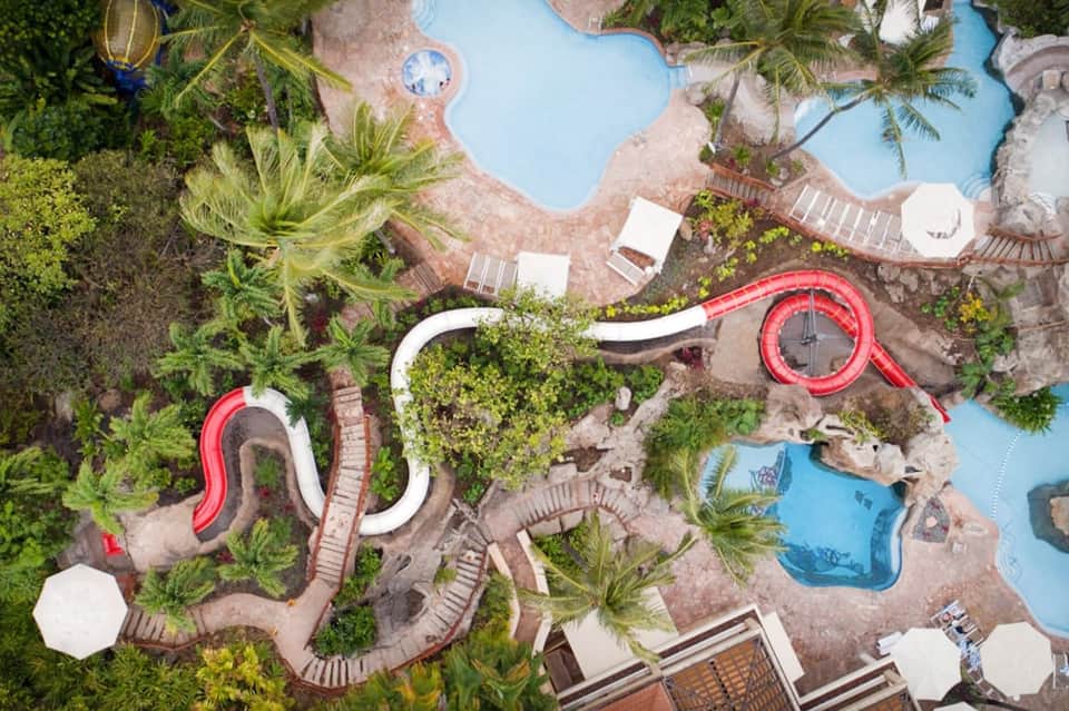 Aerial view of resort pools with red water slides, palm trees, and lounge areas