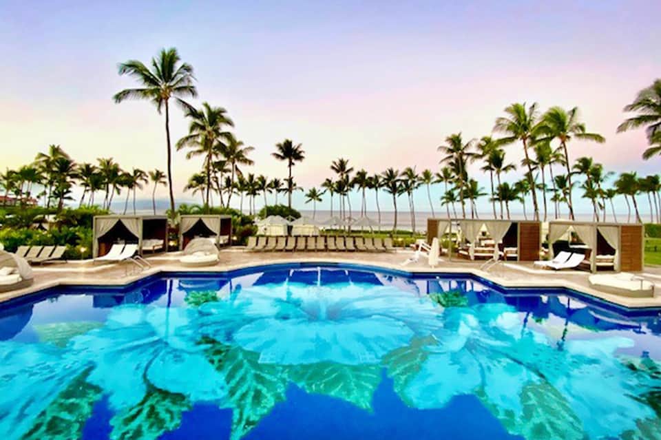Resort pool with ocean view at dusk, surrounded by palm trees and cabana seating areas