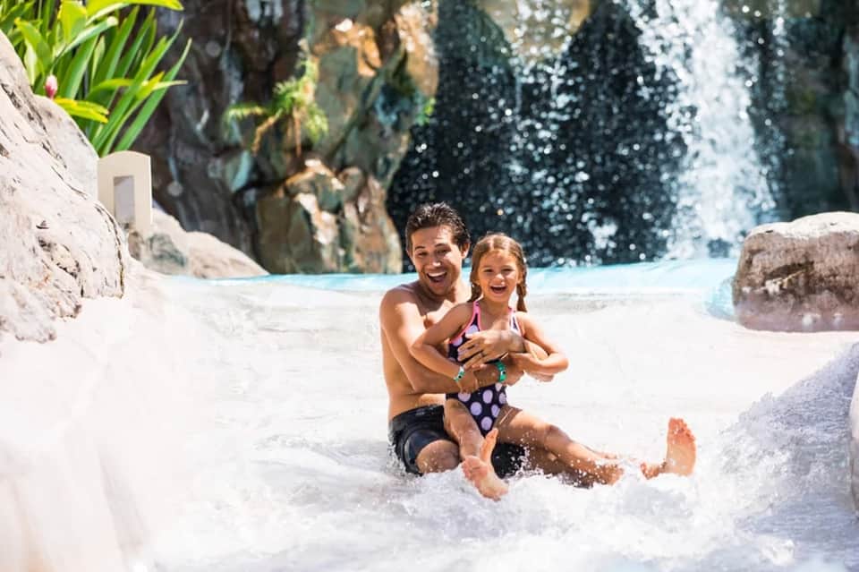 Father and daughter playing in shallow resort pool with waterfall and rock features