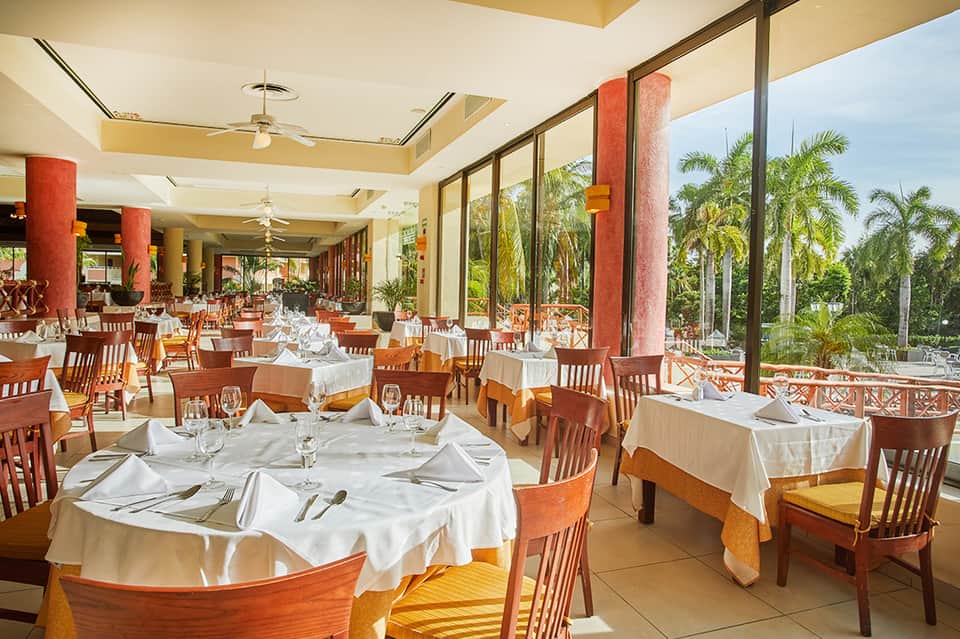 Upscale dining room with white tablecloths, wooden chairs, red columns, and tropical garden views through large windows