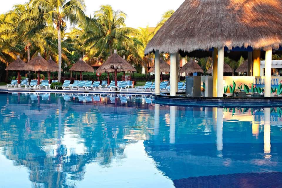 Large resort pool with thatched umbrellas, lounge chairs, palm trees, and pavilion