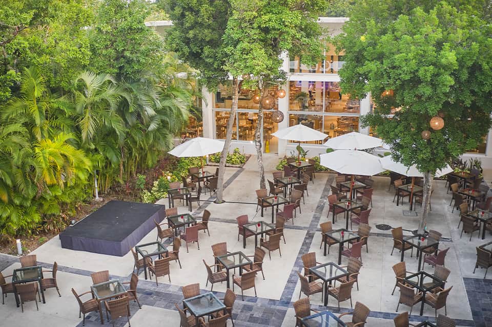 Tropical courtyard with shaded dining area, umbrella tables, green foliage, and modern building