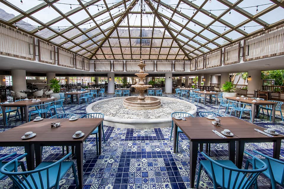 Elegant dining hall with blue chairs, central fountain, patterned tile floor, and glass ceiling
