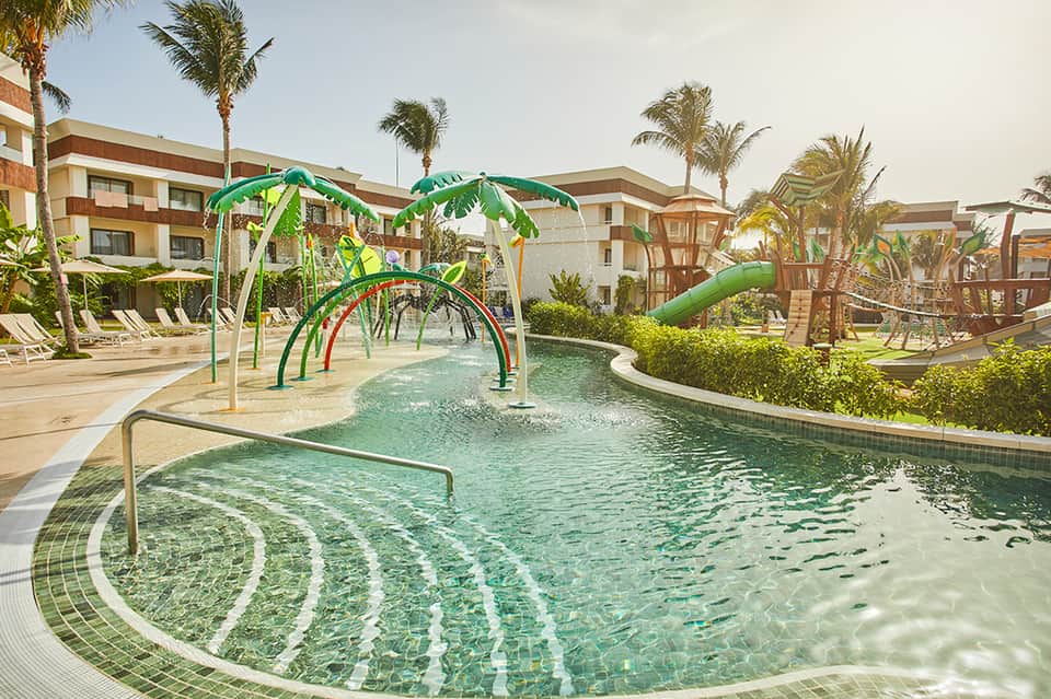 Family-friendly splash pad with green water play structures and palm-tree-lined resort buildings