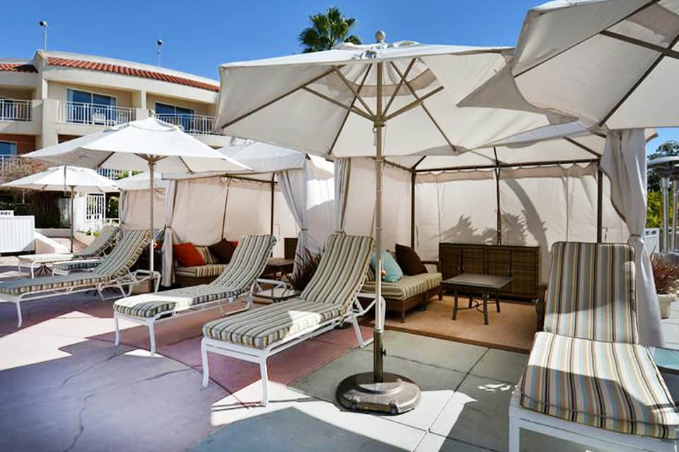 Shaded poolside lounge area with striped loungers, white umbrellas, and residential buildings