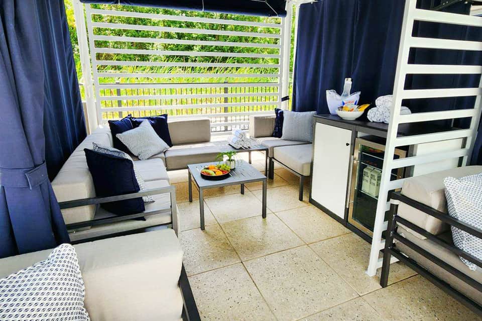 Covered outdoor lounge area with navy curtains, cream cushioned seating, striped fence view of greenery, and modern furnishings