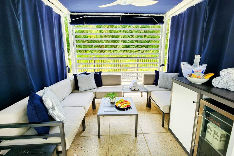 Modern cabana lounge with white sectional sofa, blue curtains, kitchenette, and lush garden view