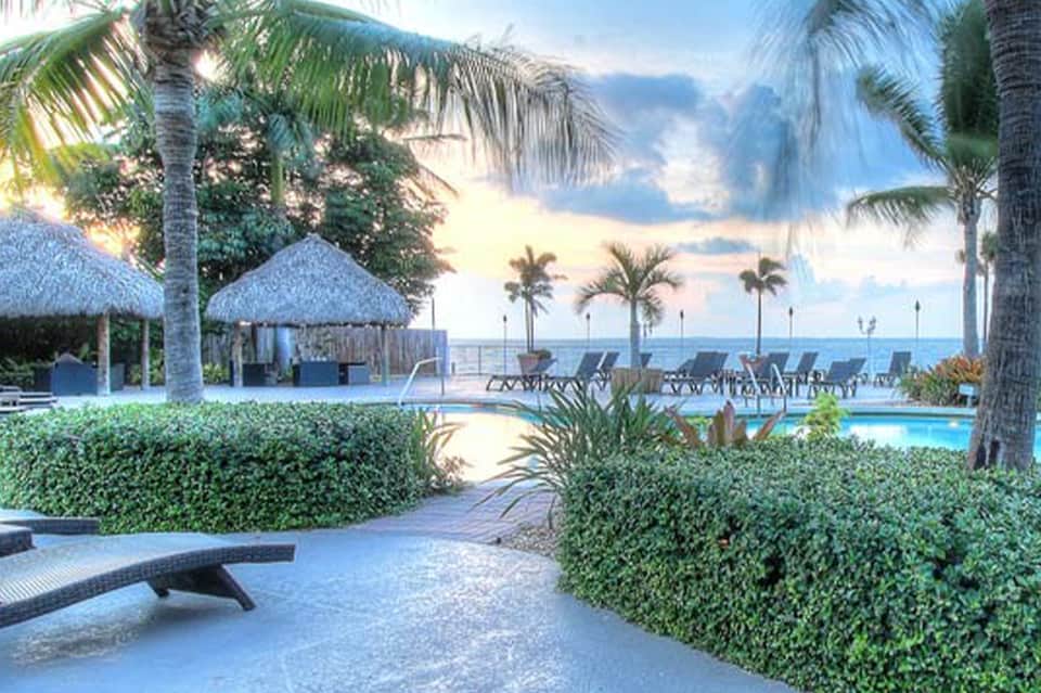 Resort pool area with thatched-roof cabanas, palm trees, lounge chairs, and ocean view at sunset