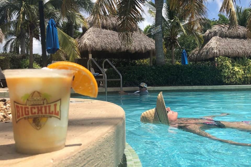 Tropical pool with tiki bar drink in foreground, thatched umbrellas, and palm trees