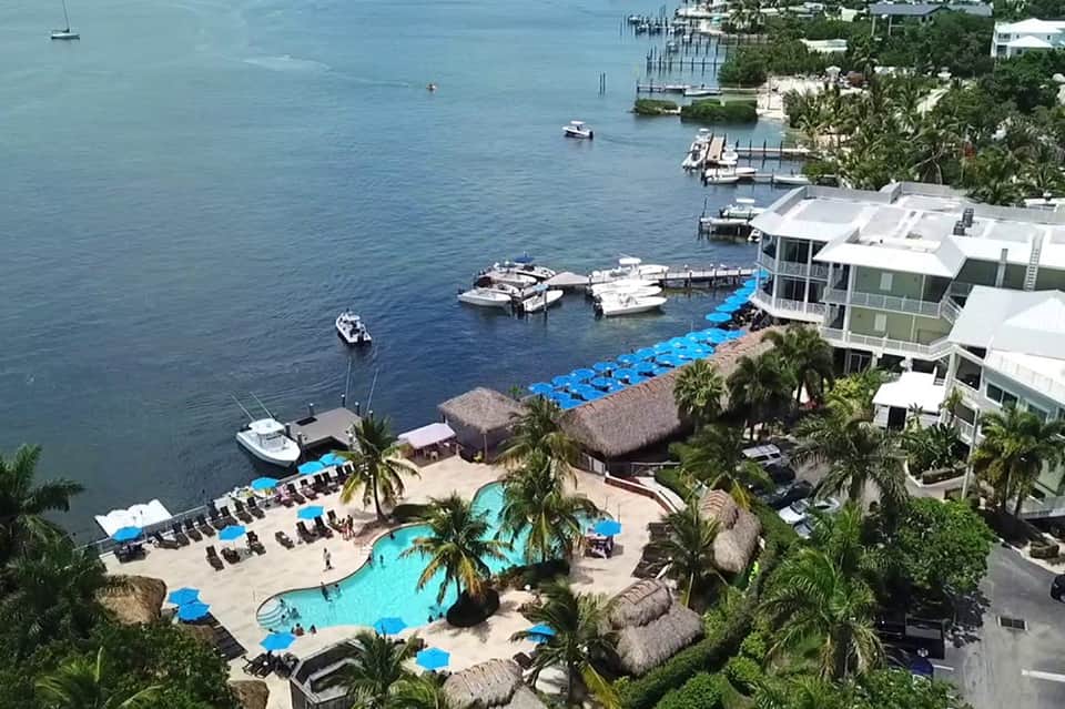 Aerial view of waterfront resort with turquoise pool, beach, boats, and palm-lined grounds