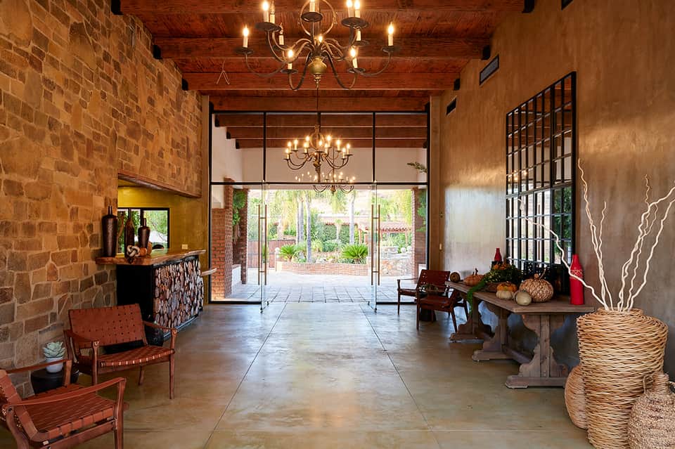 Rustic lodge entryway with exposed wood beams, stone fireplace, chandelier, and glass doors opening to courtyard