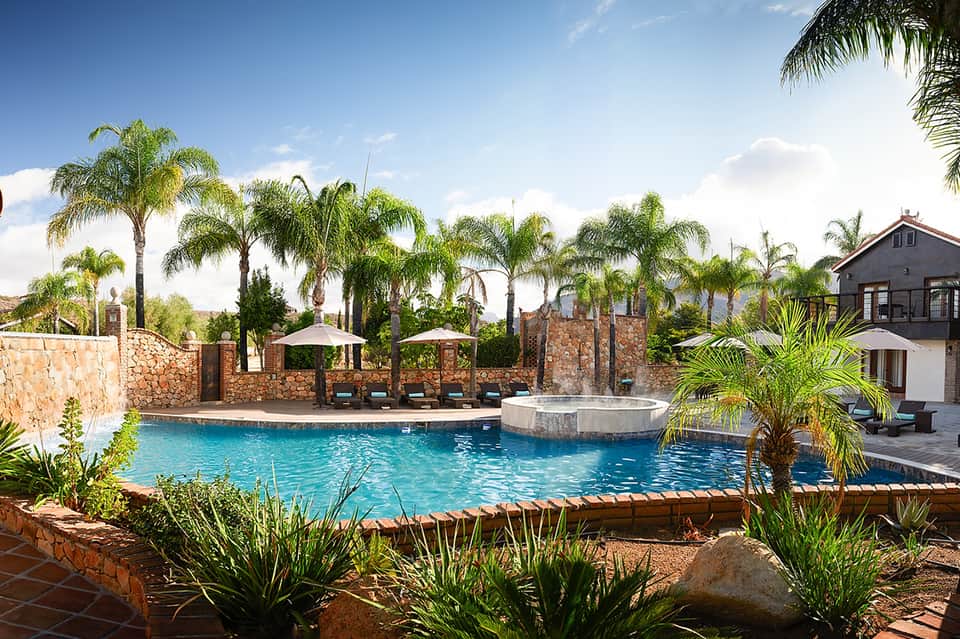 Resort pool with stone wall, palm trees, umbrellas, and Mediterranean-style building with mountain views