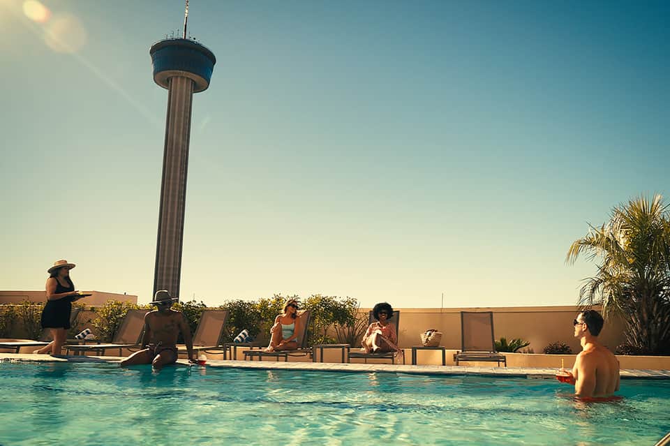 Resort rooftop pool with loungers, guests swimming, observation tower visible, palm trees, and clear blue sky