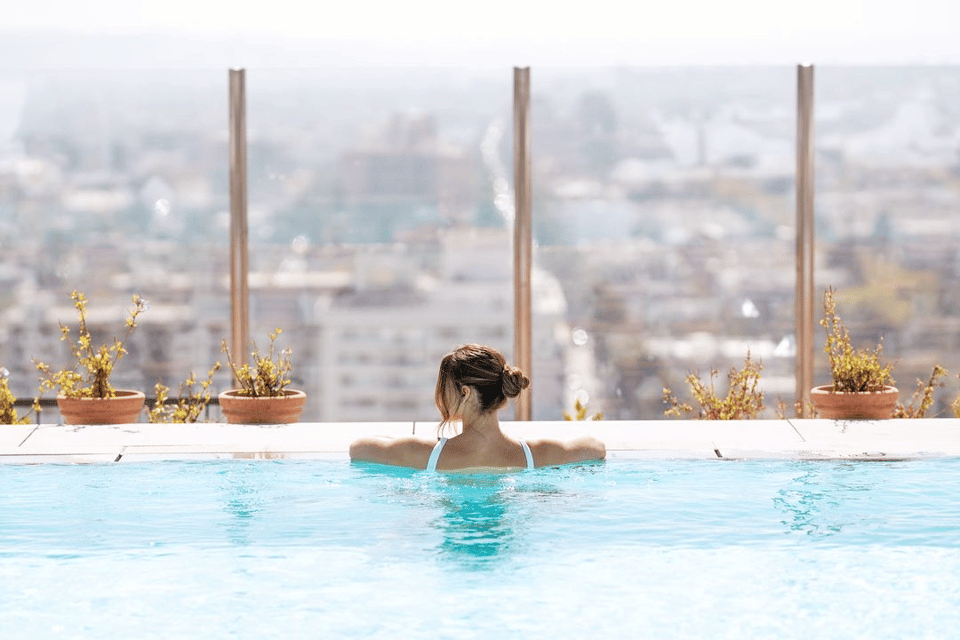 Woman in rooftop infinity pool with cityscape view and potted plants in background