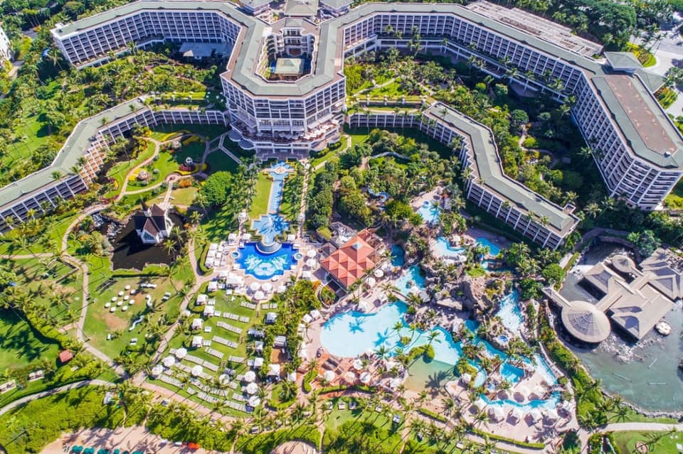 Aerial resort view showing multiple curved buildings with solar panels, lush gardens, and multiple pools