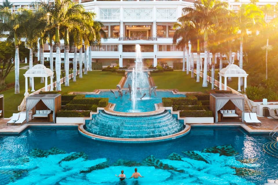 Grand resort courtyard with central fountain, white gazebos, palm trees, and lagoon-style pool at sunset