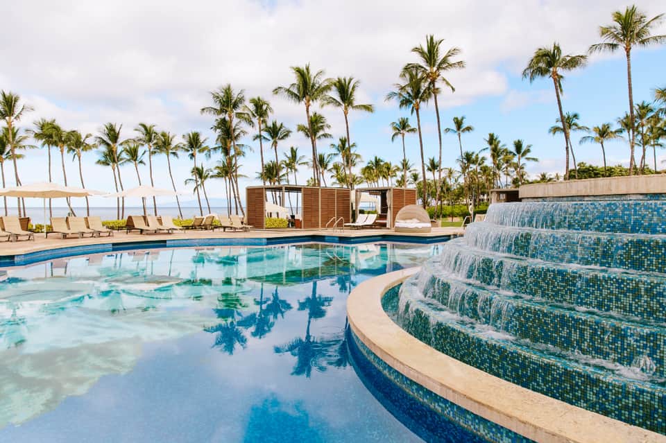 Modern resort pool with waterfall feature, lounge chairs, palm trees, and ocean views