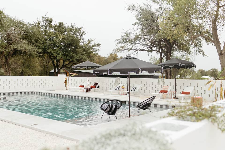 Resort pool with white privacy fence, black umbrellas, loungers, and mature trees surrounding