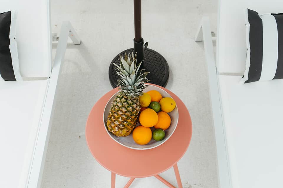 Overhead view of coral table with pineapple and citrus fruits between white lounge chairs
