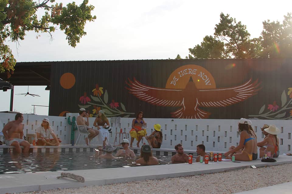 Community pool with white fence, people swimming, mural backdrop, and summer gathering area