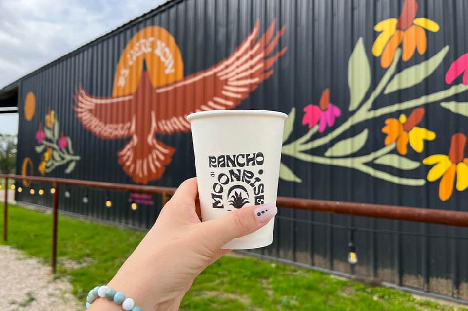 Hand holding Rancho Moneisa cup in front of colorful mural with bird and flower designs