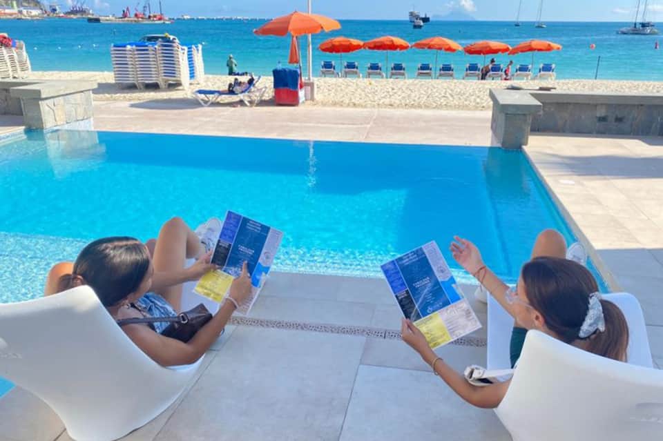 Resort beachfront pool with ocean view, lounge chairs, orange umbrellas, and sandy beach with umbrellas beyond