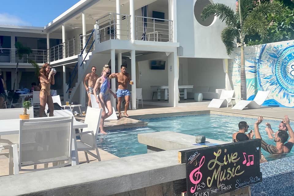 Modern resort pool with guests, white multi-level building, palm trees, and live music event