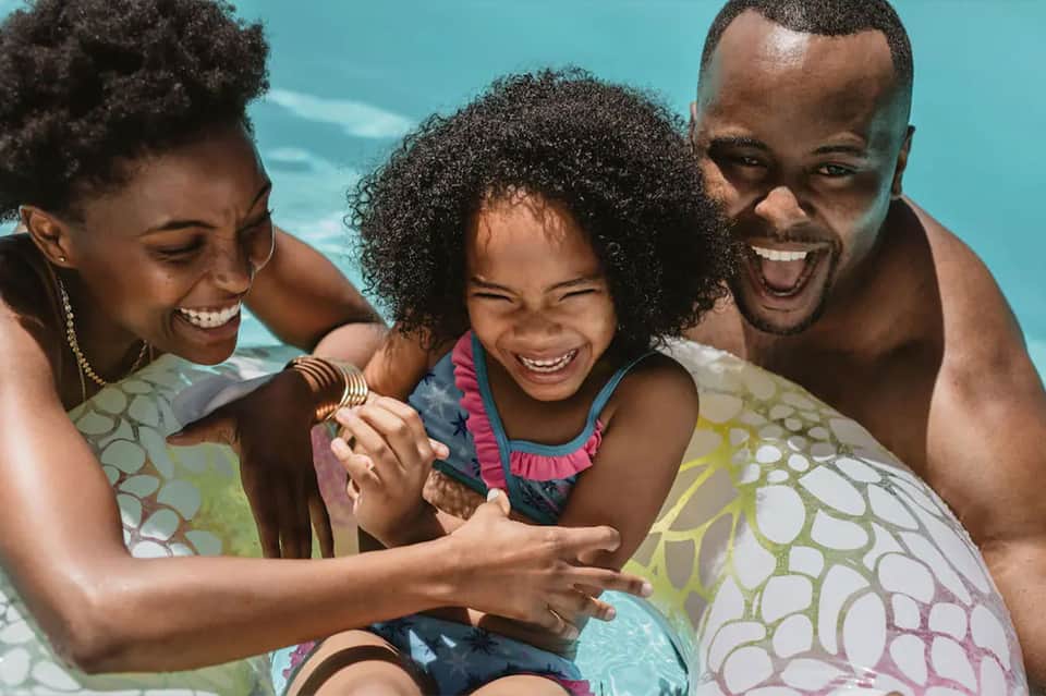 Family of three smiling and playing together in shallow turquoise ocean water under bright sunshine